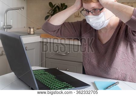 A Middle-aged Woman In A Medical Mask And Glasses For A Laptop In Cozy Kitchen Expresses Emotions Of