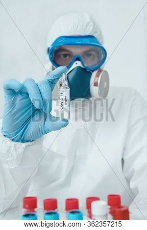COVID-19 or 2019-nCoV. Doctor microbiologist scientist in a protective PPE suit shows the ampoule with a vaccine against coronavirus. Vaccination.