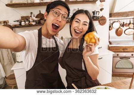 Image of multicultural excoted couple taking selfie on smartphone and smiling while holding bell pepper in cozy kitchen