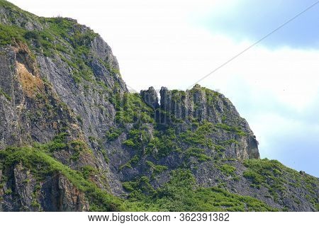 A Fragment Of A Mountain Range Overgrown With Green Shrubs And Grass, View Against A Blue Cloudy Sky