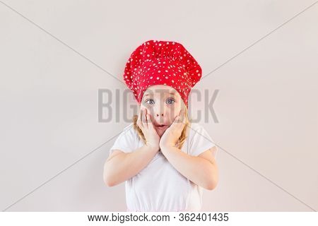 Little Blonde Smiling Girl In Red Cooks Hat Staying On Neutral Grey Background And Holding Her Cheek