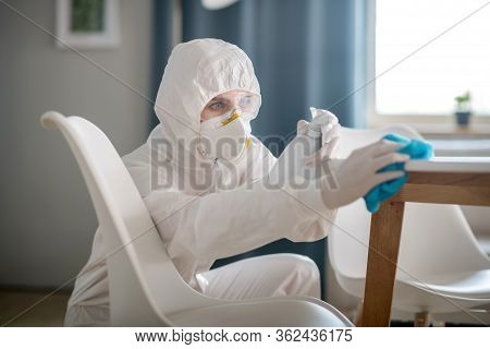 Woman In White Workwear And Protective Gloves Sparying The Antiseptic Liquid On The Furniture