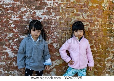 Asian Identical Female Twins Child Wearing Jacket During Cold Weather And Standing On An Old Brick W