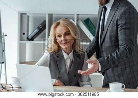 Businessman Pointing With Hand Near Smiling Businesswoman Looking At Laptop On Table In Office
