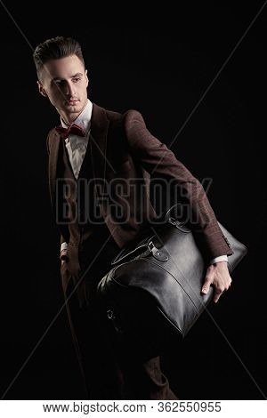 Portrait of a handsome man in elegant classic suit and bow tie posing with leather bag on a black background. Business style. Men's fashion, accessories.