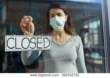 small business, pandemic and service concept - young woman in protective medical face mask hanging banner with closed word on door or window
