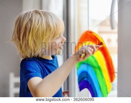 Little Boy Sitting At Window With Drawing Rainbow While Coronavirus Quarantine. Rainbow Sign Is Symb