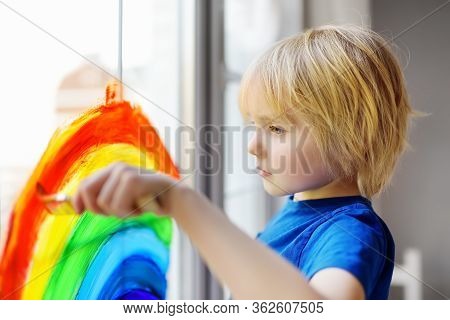 Little Boy Sitting At Window With Drawing Rainbow While Coronavirus Quarantine. Rainbow Sign Is Symb