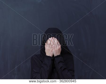 portrait of beautiful muslim woman in fashionable dress with hijab making traditional prayer to God, keeps hands in praying gesture isolated on black chalkboard background representing modern islam 