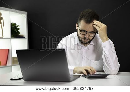 Young Handsome Puzzled Businessman In White Shirt And Glasses Sitting At Desk With Computer, Looks S