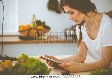 Young Woman Looking For A New Recipe For Cooking In A Kitchen. Housewife Is Making Online Shopping B