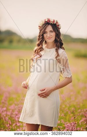 Portrait Of A Pregnant Woman In Fieid Of Pink Flowers. Young Beautiful Pregnant Girl With A Wreath O