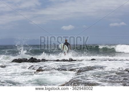 The Surf Enters The Water. Male Surfer Entering The Sea With His Board In A Black Surfing Suit. Tene