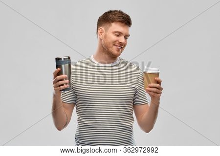 eco living and sustainability concept - happy smiling young man comparing thermo cup or tumbler with disposable paper coffee cup over grey background