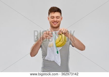 food, sustainability and eco living concept - smiling young man in striped t-shirt putting bananas into reusable string bag over grey background