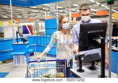 shopping, sale, consumerism and people concept - happy couple in face protective medical masks for protection from virus disease buying food at supermarket self-service cash register