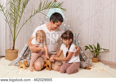Horizontal Shot Of Handsome Man Wearing White T Shirt Sitting On Floor With Her Daughters And Having