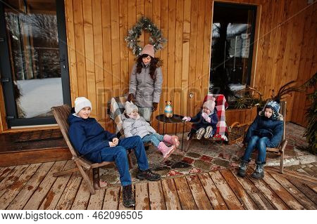 Attractive Mother With Her Four Kids On The Terrace Of A Wooden House  In Winter Day.