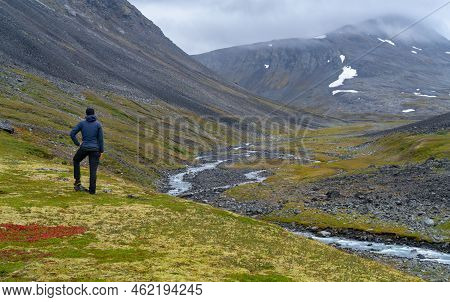 Female Hiker Overlooking Wild River In Harsh, Remote Arctic Valley On A Cloudy, Rainy Day Of Summer.