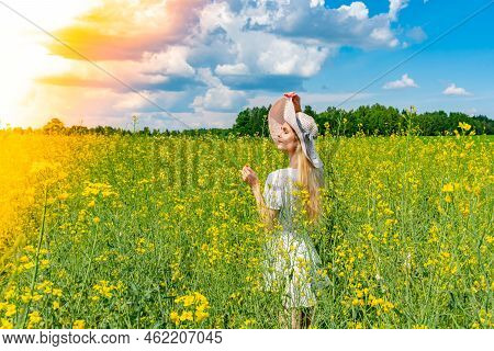 Beautiful Girl In Wide-brimmed Hat In A Flowering Field With White Dress. Sunbeams, Glare, Light Lea