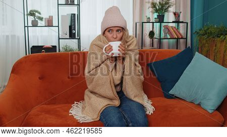 Sick Girl Wear Hat Wrapped In Plaid Sit Alone Shivering From Cold On Sofa Drinking Hot Tea In Unheat