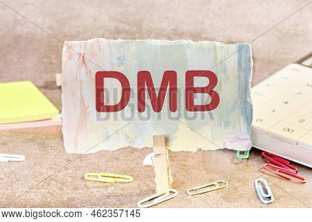 Dmb Text On A Piece Of Colored Paper Held By A Clothespin Next To Office Supplies.
