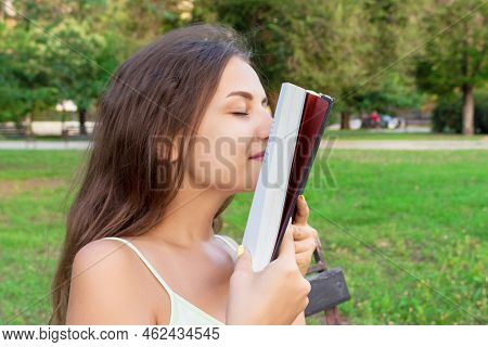 Beautiful Young Brownhair Girl Smells A Fresh Printed Book Sitting On The Bench In The Park.
