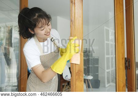 Beautiful Young Asian Woman Cleaning Window At Home Using Window Spray And Wearing Yellow Gloves. Wo