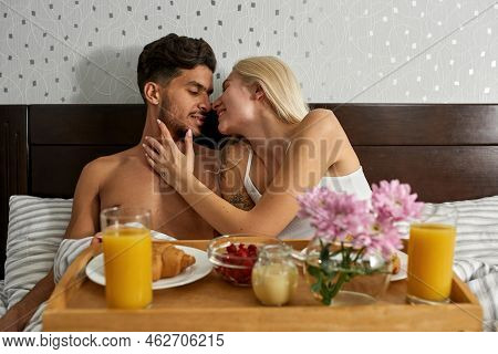 Young Plesed Mutiethnic Couple Resting And Having Breakfast On Bed At Home. Caucasian Woman Touch An