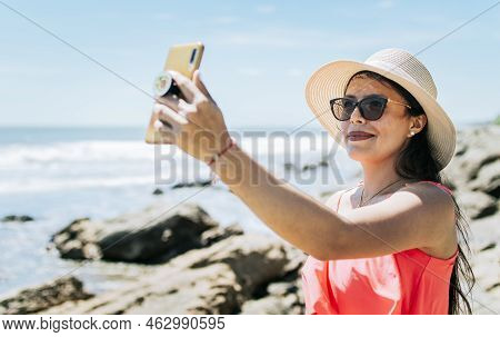 Girl In Hat On The Beach Taking A Selfie. Young Woman On Vacation Taking Photos On The Beach, Smilin