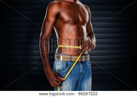 Mid section of a fit shirtless man measuring waist against black background