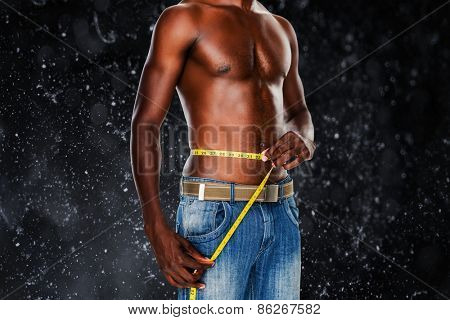 Mid section of a fit shirtless man measuring waist against black background