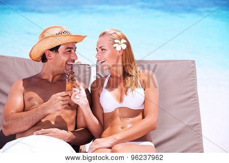 Happy couple on the beach sitting on sunbed and eating tasty sweet ice cream, with love looking on each other, enjoying romantic summer vacation 