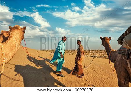 Rajasthan travel background - two indian cameleers (camel drivers) with camels in dunes of Thar desert. Jaisalmer, Rajasthan, India