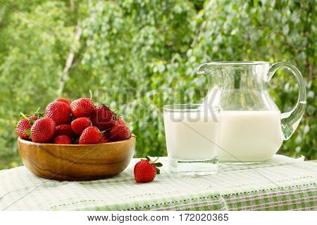 Strawberries in a wooden bowl, a glass  of milk and a jug of milk. On a table, on background of green trees.
