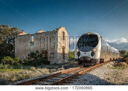 Train passing a derelict station at Lumio in the Balagne region of Corsica with snow covered mountains in the distance under a deep blue sky