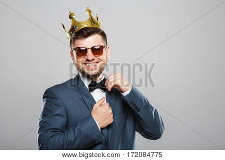 Stylish young man in suit with bow and sunglasses. Wearing crown. Looking at camera, touching bow and smiling broadly. Outrageous, fancy look, cool. Waist up, studio, indoors