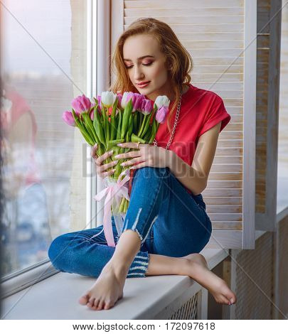 young attractive girl in blue jeans sitting with bouquet of tulips on window-sill