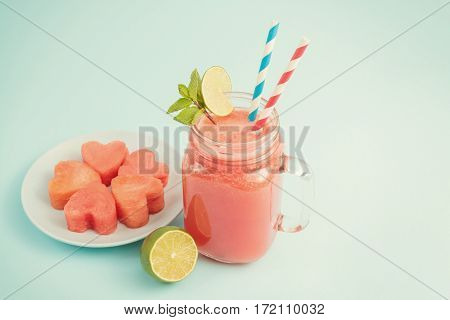 Watermelon smoothie in Mason jar decorated with lime, mint, straws with copyspace. Watermelon slices curved like hearts on plate on blue background