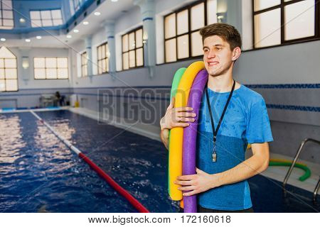 Fit smiling man holding foam roller at swimming pool. Young swimming instructor