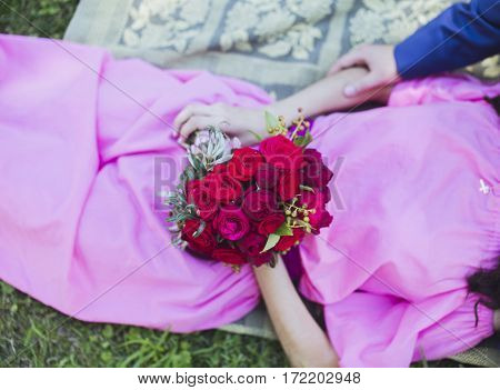 Happy couple in love lying in green grass on their wedding day. Bride in simple stylish pink dress holds beautiful wedding bouquet nice flowers. Happy people on summer day outdoors.