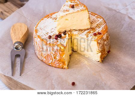 Top view of cut slice of French or German soft cheese with orange rind with mold creamy texture red pepper corns forkon parchment paper wood cutting board