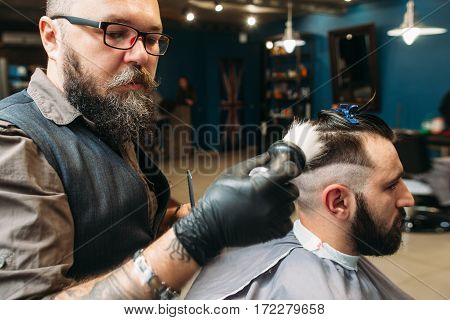 Stylist shaving client head side view. Stylish bearded barber making hairstyle for youn man in barbershop interior. Beauty, modern lifestyle, style, concept