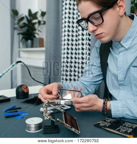 Serious young man fixing mobile phone free space. Attentive geek in glasses examing broken smartphone, repair shop workplace with tools. Electronic fixing, modern technology, business concept