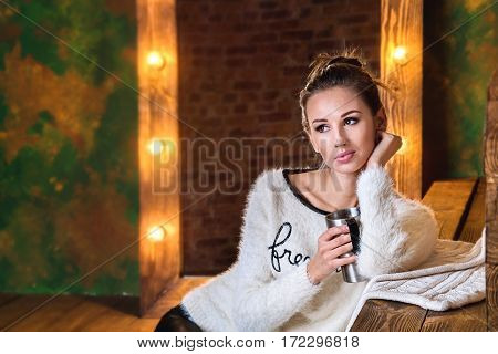 Young brunette holdind a thermocup, sitting in front of mirror with lights and looking on a side