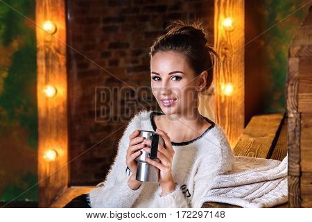 Young brunette holdind a thermocup, sitting in front of mirror with lights and looking on camera