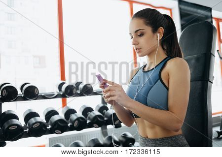 Portrait Of Young And Attractive Woman In Blue Top And With Ponytail Standing In Gym, Using Her Mobi