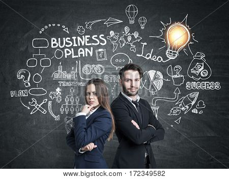 Portrait of a businessman with crossed arms and his female business partner standing near a blackboard with a business plan skech drawn on it.