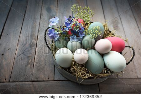 Pastel colored Easter eggs with flowers in a bowl of straw on a dark rustic wooden table