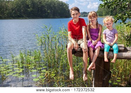 Three children of different age - teenager boy, elementary age girl and toddler girl sitting on a wooden pier by a forest lake looking at camera smiling